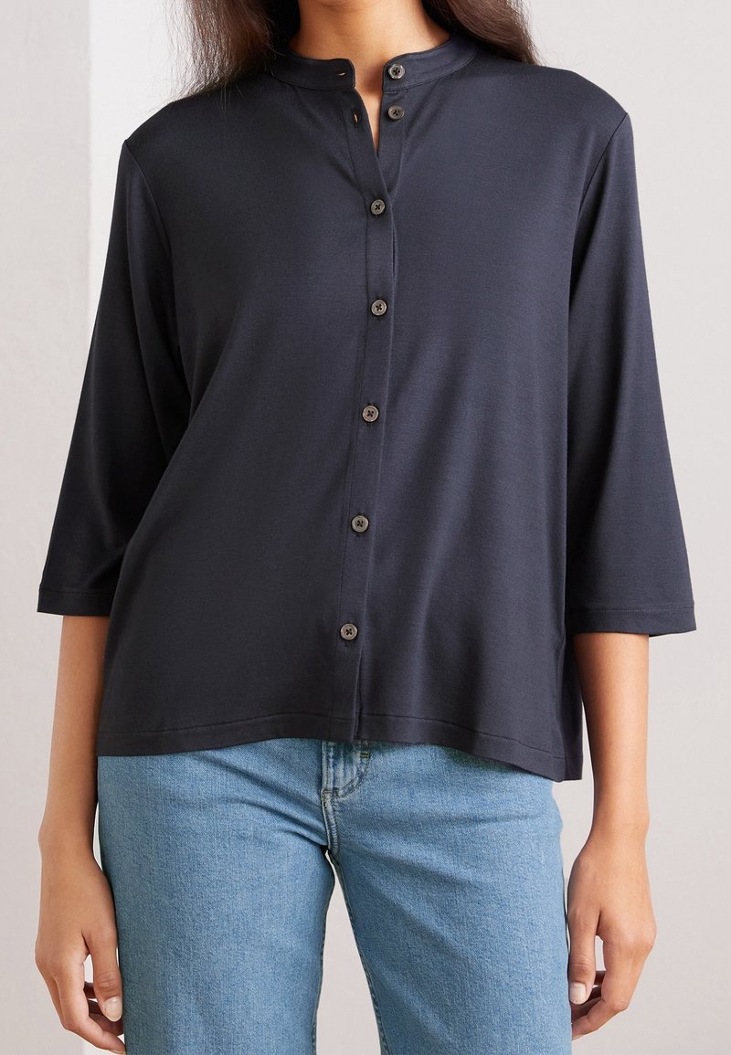 Femme portant une chemise noire à manches longues boutonnée avec un col mandarin et un jean en denim bleu clair, les bras détendus le long du corps.