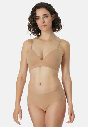 Femme aux cheveux bouclés foncés portant un soutien-gorge nude sans coutures et un slip taille haute, debout devant un fond clair uni.