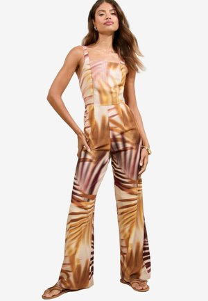 Modella posa con sicurezza in una tuta floreale con vivaci motivi a palme, mostrando un look elegante e estivo con sandali.