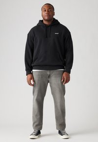 Hombre de pie vistiendo un hoodie negro de Levi's, jeans grises y zapatillas Converse negras frente a un fondo gris liso.