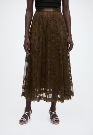 Femme portant une jupe midi en dentelle florale marron avec une ceinture unie, associée à des sandales à talons argentées à brides sur fond blanc.