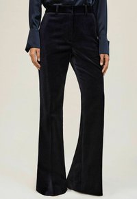 Vrouw in hoge, brede zwarte fluwelen broek met een donkerblauwe satijnen blouse met lange mouwen.