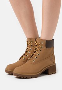 Bottines en cuir beige avec un accent marron au col, œillets dorés, lacets jaunes et rouges, et un talon épais et texturé.