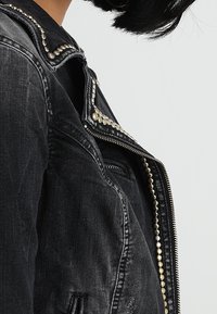 Osoba v černé džínové bundě se stříbrnými kolíky podél švů a zipu, ukazující částečný boční profil a černé vlasy.