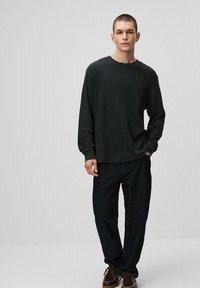 Ciemnozielona koszula z długim rękawem o luźnym kroju i prążkowanej fakturze, zestawiona z ciemnoniebieskimi spodniami dżinsowymi oraz brązowymi sznurowanymi butami.