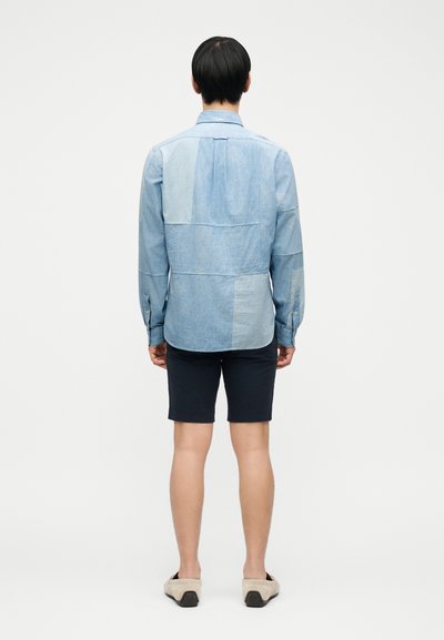 Άνδρας, γυρισμένος πλάτη, φορώντας ένα μπλε τζιν πουκάμισο πατς, μαύρα σορτς και μπεζ παπούτσια χωρίς κορδόνια, σε απλό φόντο.