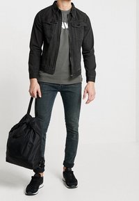 Giacca di jeans nera con due tasche sul petto sopra una t-shirt grigia con testo bianco, jeans scuri, sneaker nere e una borsa nera con cordoncino.