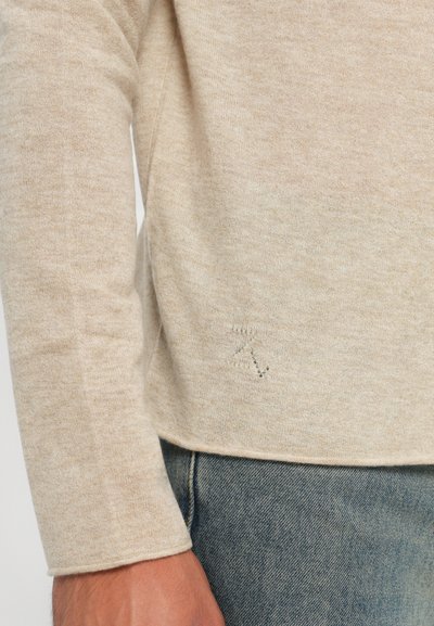 Vaaleanbeige neule, joka on valmistettu pehmeästä, tekstuurilla varustetusta kankaasta. Siinä on pitkät hihat ja vasemmalla puolella on hillitty "Z"-kuvio. Yhdistetty farkkuihin.