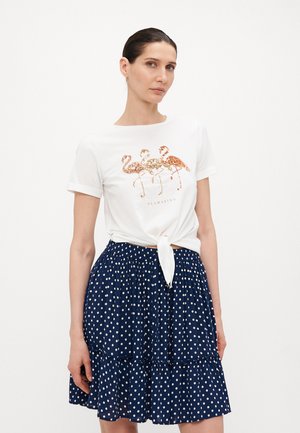 Hvit t-skjorte med kort erm og en paljett flamingo-grafikk, knyttet i livet. Kombinert med et marineblått prikkete skjørt med lagdelt design.