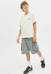 T-shirt jaune clair avec le texte "ORIGINALS", associé à un short cargo gris avec de grandes poches, des chaussettes blanches et des baskets noires.