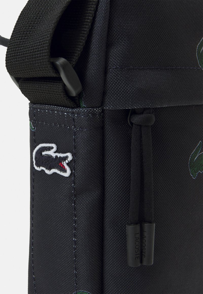 Sac à épaule en tissu noir présentant un logo crocodile brodé, une texture particulière et une sangle ajustable avec des accents en métal.