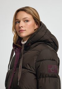 Brun puffjacka med huva, med vinröda detaljer, en logopatch och dragsnören. Slät, vadderad textur med dragkedjestängning.