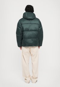 Dunkelgrüne Pufferjacke mit Kapuze, charakterisiert durch horizontale Nähte, eine glatte Textur und eine lockere Passform, kombiniert mit hellbeigen Hosen.