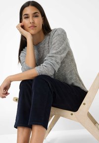 Jeune femme avec de longs cheveux noirs portant un pull gris et un pantalon sombre, assise sur une chaise en bois, reposant la tête sur sa main, avec une expression détendue.
