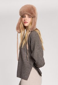 Brączowa dzianinowa czapka z pluszowym, jasnobrunatnym futerkiem w środku, klapkami na uszy i wiązaniami. W połączeniu z ciemnoszarym swetrem w prążki i jasnobeżowymi spodniami.