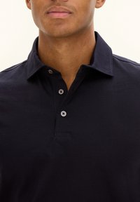 Navy blue polo shirt made of cotton, featuring a collared neck, three-button placket, and textured fabric, set against a neutral background.
