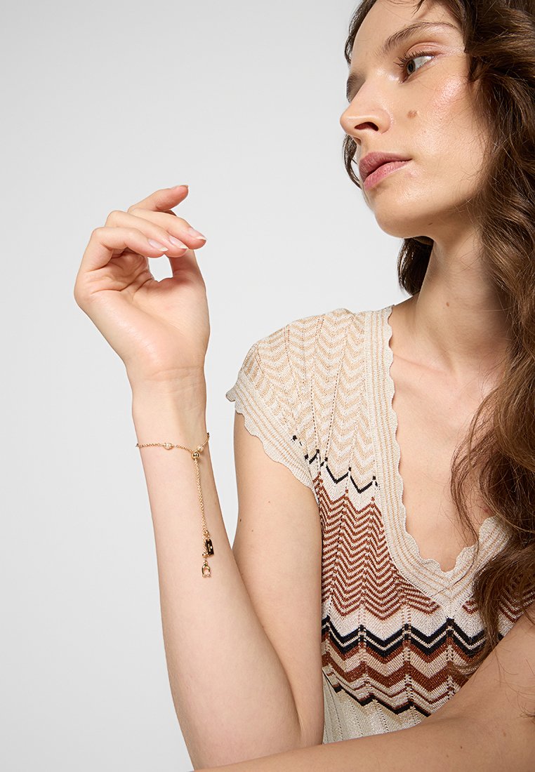 Bracelet en or avec une chaîne délicate, comportant un petit charm et un élément pendentif. Porté au poignet d'un modèle, mettant en valeur un design complexe.