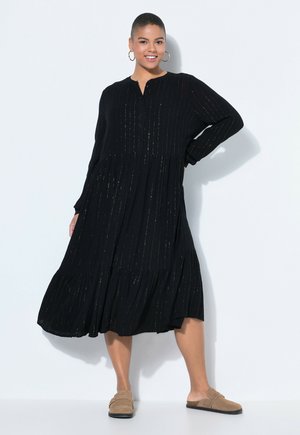 Uśmiechnięta kobieta w czarnej, długorękawowej sukience midi z delikatnymi paskami, kolczykami kołami i beżowymi butami wsuwanymi, pozująca na białym tle.