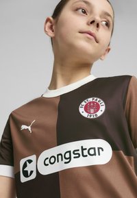 Brūna un tumši brūna futbola krekls ar īsām piedurknēm, ar baltu apkakli, Puma logotipu, FC St. Pauli emblēmu un "congstar" logotipu.