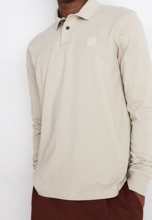 Mand iført beige langærmet poloshirt med en knap lukket og brune bukser, stående mod en hvid baggrund.