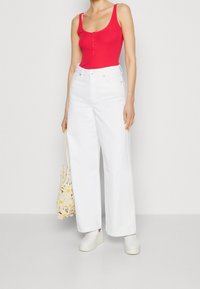 Kvinna klädd i en ljusröd ärmlös topp, hög midja vid ben-bukser med vita byxor, vita slip-on skor, hållande en krämfärgad virkad tote-väska med gula blommor.