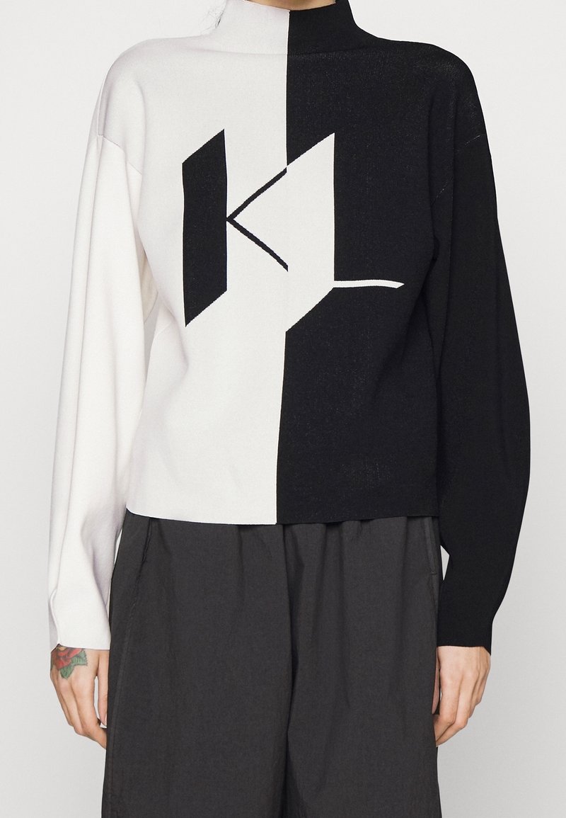 Pull à manches longues noir et blanc avec un col montant et un motif géométrique abstrait coupé au milieu, porté avec un pantalon large foncé.