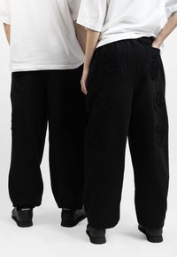 Black sweatpants with textured patches in various shapes. Loose fit with elastic waistband, paired with white oversized t-shirts and black shoes.