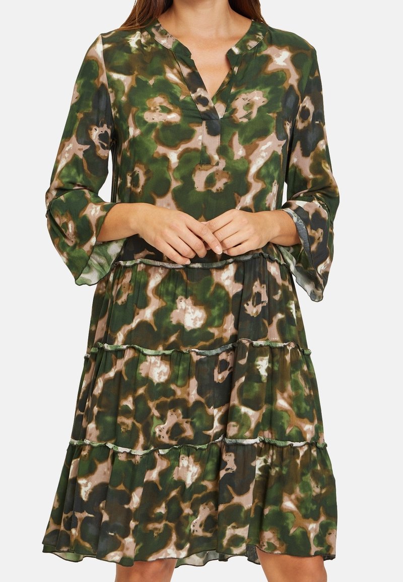 Femme portant une robe à motifs floraux verts et bruns, manches 3/4, jupe à volants et décolleté en V, mains jointes à la taille.