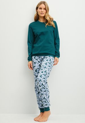 Femme debout pieds nus, portant un haut à manches longues bleu sarcelle et un pantalon de jogging à motifs floraux sur un fond clair uni.