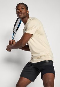 Μπεζ αθλητική μπλούζα και μαύρη βερμούδα, λεία υφή, εφαρμοστή σχεδίαση, διακοσμητικά λογότυπα, κρατώντας ένα τενιστικό ρακέτα, ουδέτερο φόντο.