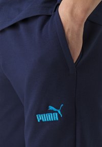 Navy blue athletic pants made of soft fabric with a side pocket and a light blue Puma logo embroidered on the thigh.