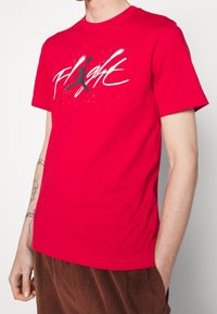 Punainen puuvillainen T-paita, jossa on suuri musta "flight" -grafiikka roiske-yksityiskohdilla. Lyhyet hihat ja pyöreä kaula-aukko.