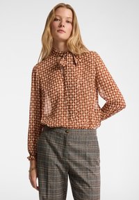 Braun gemusterte Bluse mit einem Bindekragen, aus einem durchsichtigen, leichten Material. Kombiniert mit grauen Karo-Hosen, die eine taillierte Passform zeigen.
