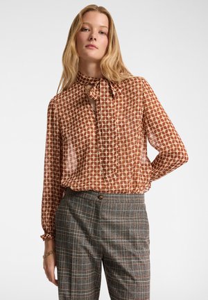 Bruine blouse met patroon en striknek, vervaardigd uit een doorschijnende, lichte stof. Gecombineerd met grijs geruite broek met een getailleerde pasvorm.