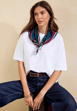 Jonge vrouw met lang bruin haar, draagt een wit T-shirt, donkere jeans, een blauw-paarse patroon sjaal, gouden oorbellen en armbanden, zittend.
