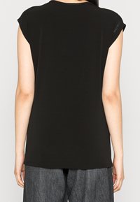 Žena v bezramínkovém černém topu s malým logem Calvin Klein na rameni, v kombinaci s volnými šedými kalhotami, zachycena zezadu.