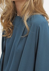 Turkusowa bluzka wykonana z lekkiego materiału, charakteryzująca się luźnym krojem, subtelnym drapowaniem i okrągłym dekoltem. Prosty design z minimalną ilością detali.