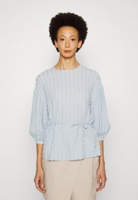Blusa azul clara com tecido texturizado, decote redondo e mangas bufantes. Apresenta um laço na cintura e padrão de riscas verticais.