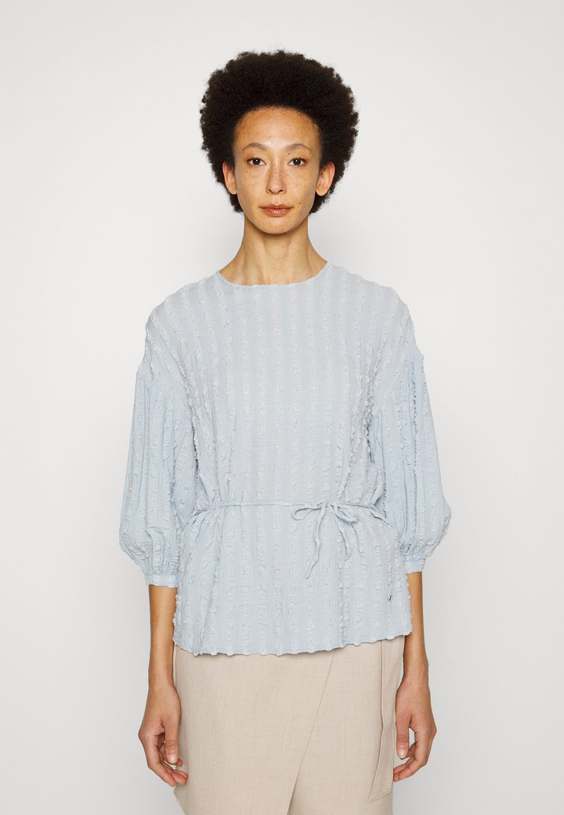 Blusa azul clara com tecido texturizado, decote redondo e mangas bufantes. Apresenta um laço na cintura e padrão de riscas verticais.