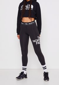 Mustat lyhytcroppinen huppari, jossa on valkoinen logo, yhdistettynä mustiin legginsseihin, joissa on valkoista tekstiä, ja mustiin lenkkitossuihin, joissa on mustavalkoiset raidalliset sukat.