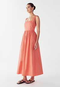 Maksi haljina od koralnog lana s kvadratnim izrezom, širokim naramenicama i elastičnom pojasom. Model je kombinirao s crnim ravnim sandalama.