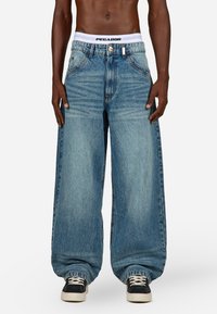 Svetlo modri preveliki kavbojke z ravno silhueto, ki imajo bel pas z blagovno znamko in diskretno zbledelost.