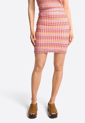 Kobieta w kolorowej szydełkowej mini spódniczce i brązowych sandałach na platformie, stojąca na tle jednolitego białego tła.