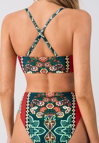 Bikini colorato con un motivo floreale geometrico. Presenta una base verde scuro, accenti rossi e spalline incrociate sul retro. Tessuto testurizzato.