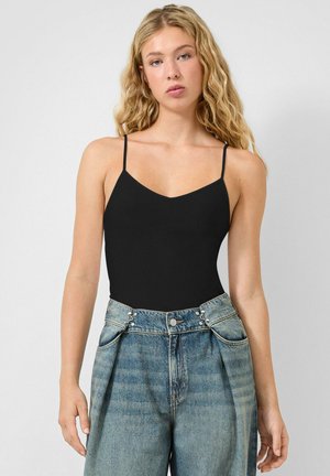 Jonge vrouw met lang golvend blond haar, gekleed in een zwart topje met spaghettibandjes en een hoge blauwe denimjeans, tegen een effen achtergrond.