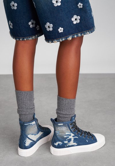 JeANSKETENI visoke z usnjenim deležem v modri barvi, bele gumijaste podplate, črne vezalke in sivi rebrasti nogavici, ki imajo cvetlične vzorce na kratkih hlačah.