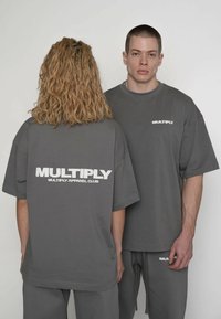 Graue, übergroße T-Shirts, präsentiert von zwei Models, mit weißem "MULTIPLY"-Schriftzug auf der Rückseite und Vorderseite, gefertigt aus weichem Baumwollstoff.