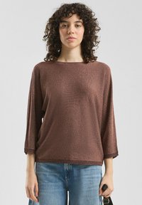 Brązowy sweter wykonany z dzianiny, z rękawami o długości trzech czwartych, szerokim dekoltem i ściśniętym dołem. Połączony z jasnoniebieskimi dżinsami.