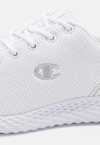 Zapato deportivo blanco con parte superior de malla, cordones redondos y suela de goma. Presenta un logo plateado en el lateral y una suela texturizada para un mejor agarre.