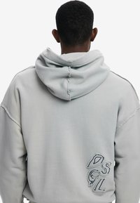 Sudadera oversized gris claro con cordón, bordes deshilachados y bordado en la parte posterior. Se puede ver la textura suave de la tela.